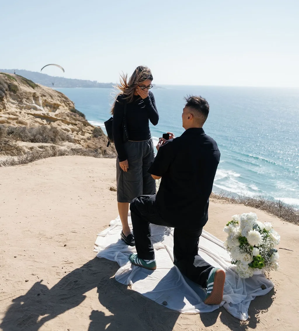 Surprise proposal at Torrey Pines Gliderport in San Diego, man on one knee with white flowers and ocean view